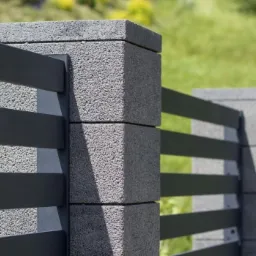 Nowoczesne ogrodzenie panelowe z szarych bloczków betonowych i poziomych, metalowych przęseł, widoczne na tle zielonego trawnika.