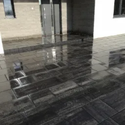 Świeżo ułożona, ciemnoszara kostka brukowa przed nowoczesnym domem z szarej cegły, odbijająca światło i tworząca lustrzany efekt.