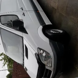 Biały samochód dostawczy marki Mercedes, widoczny z przodu pod kątem, stoi na mokrym chodniku w pochmurny dzień.