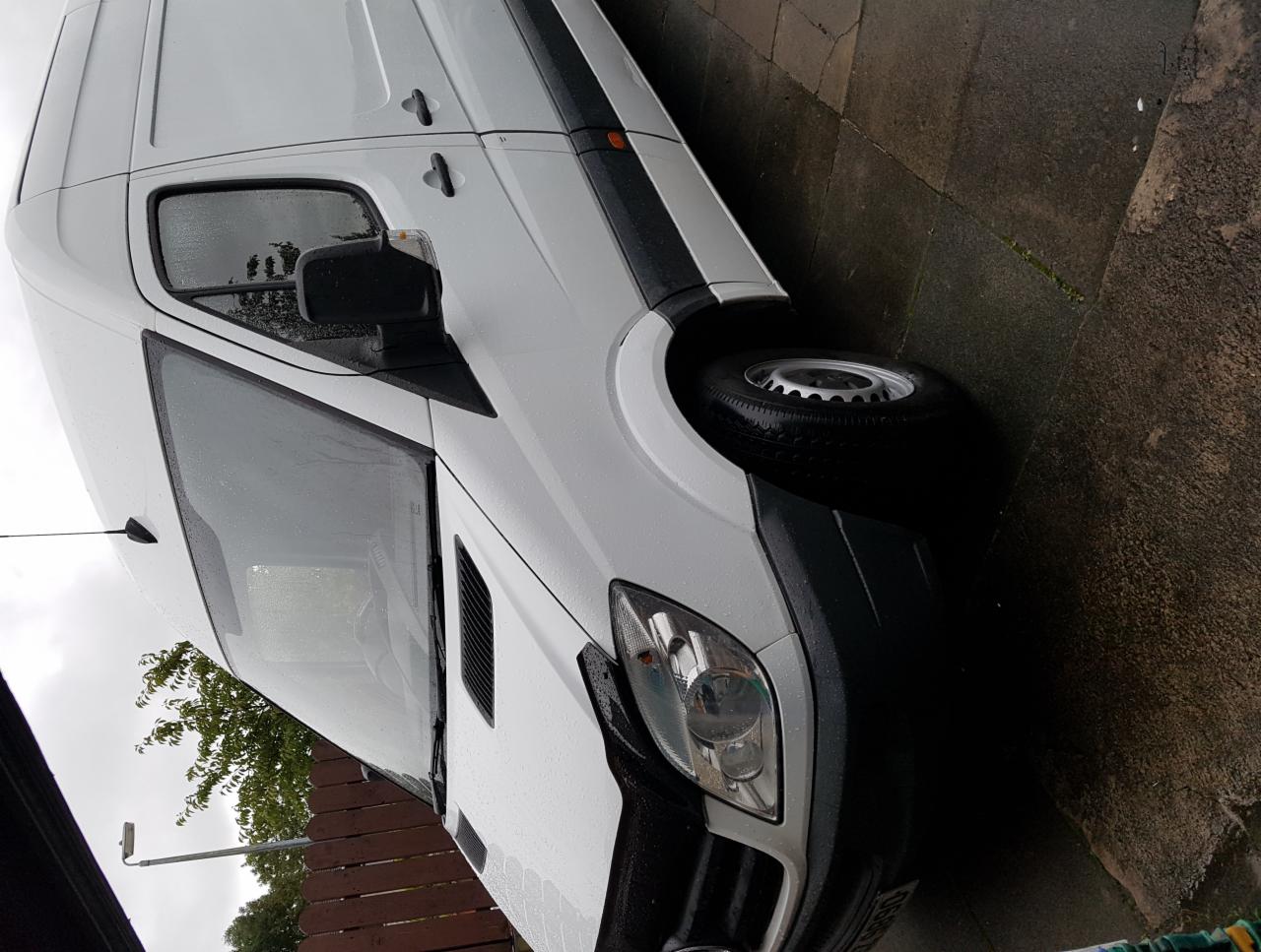 Biały samochód dostawczy marki Mercedes, widoczny z przodu pod kątem, stoi na mokrym chodniku w pochmurny dzień.