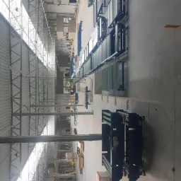 Widok z góry na długą linię transportową w hali przemysłowej z widocznymi elementami konstrukcji dachu i instalacjami pod sufitem, w tym korytami kablowymi.