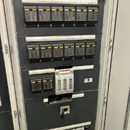 Rozdzielnica elektryczna z bezpiecznikami i wyłącznikami, widoczne oznaczenia F1-F14, Jean Muller, Apator, zainstalowana w szarej szafie metalowej.