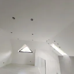 Poddasze z oknem trójkątnym i oknem dachowym, białe ściany, minimalistyczne oświetlenie punktowe, szklana balustrada na antresoli, wiszące lampy dekoracyjne.