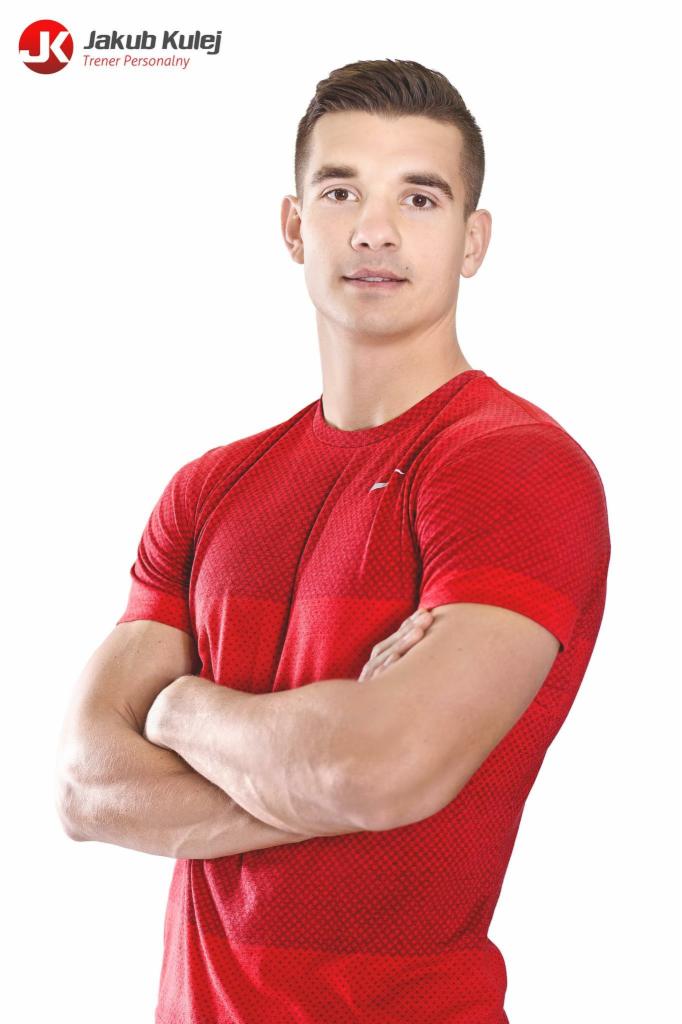 Młody mężczyzna w czerwonej koszulce sportowej z rękami skrzyżowanymi na piersiach, z logo firmy w lewym górnym rogu na białym tle.