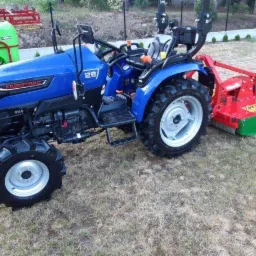 Niebieski traktor Farmtrac 26 4x4 z czerwono-zielonym glebogryzarką na trawniku. Widoczny również opryskiwacz. Sprzęt gotowy do pracy w ogrodzie.
