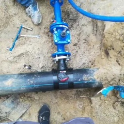 Montaż niebieskiego zaworu na czarnej rurze w wykopie. Widoczne narzędzia i fragmenty ubrań osób montujących. Piasek i ziemia w tle.