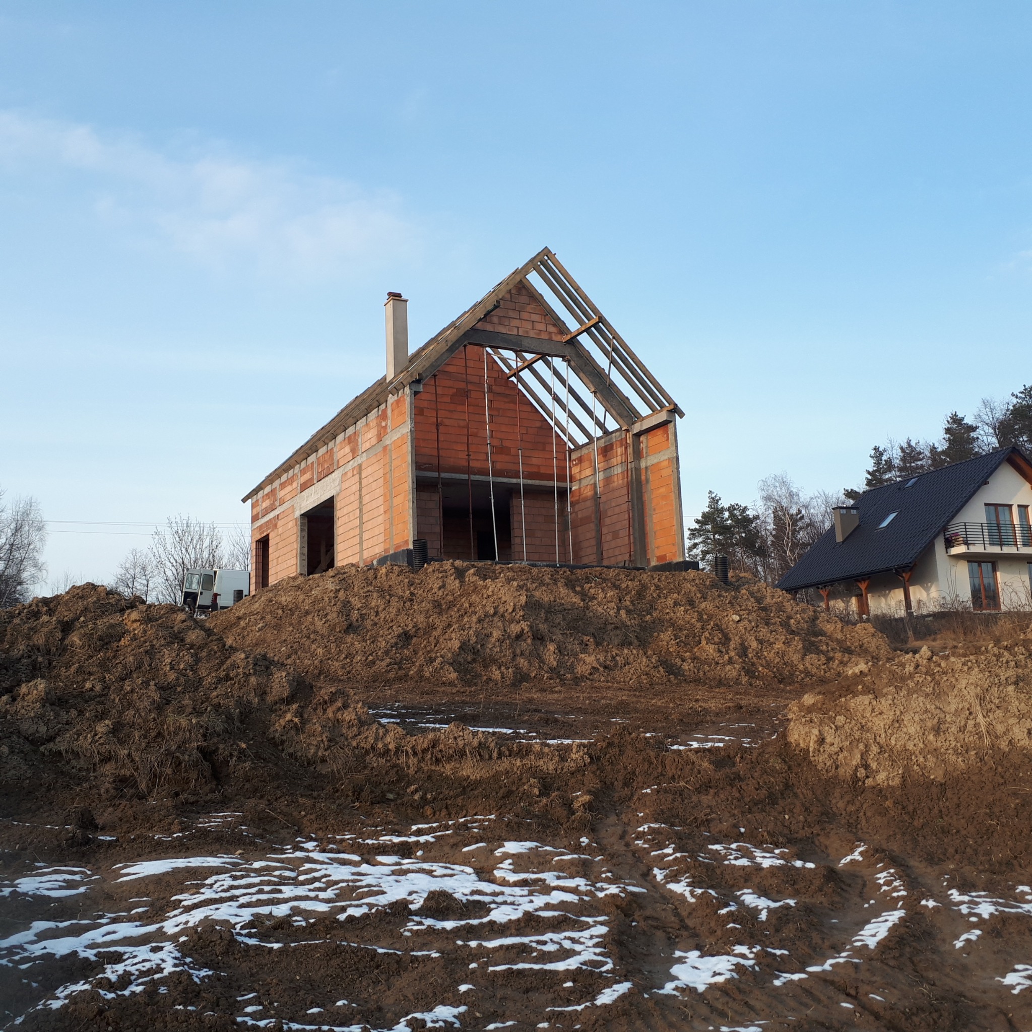 Wznoszony dom murowany z czerwonej cegły na wzniesieniu, widoczne elementy konstrukcji dachu, w tle drugi, zamieszkały dom i częściowo ośnieżona ziemia.