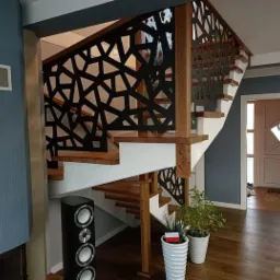 Nowoczesne schody z ażurową, geometryczną balustradą w kolorze czarnym, drewniane stopnie, białe podstopnice, widoczny głośnik i doniczki z roślinami na parkiecie.