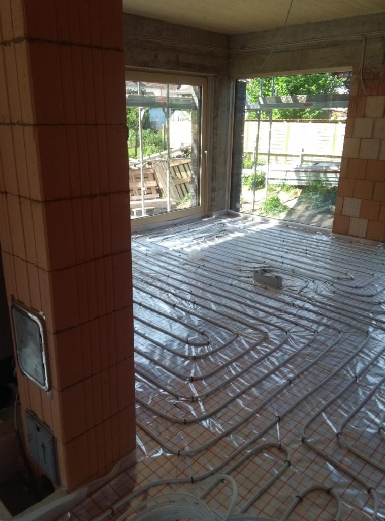 Wnętrze w stanie surowym z ułożonym systemem ogrzewania podłogowego na folii z siatką, widoczne okno z widokiem na zewnątrz i ceglany filar z metalową wnęką.