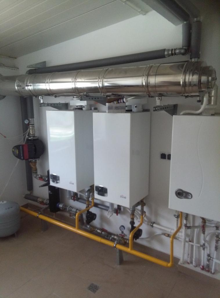 Trzy białe, wiszące kotły grzewcze marki Ferroli zamontowane na ścianie, z widocznymi rurami miedzianymi i żółtymi rurami gazowymi, oraz srebrnym kominem odprowadzającym spaliny.