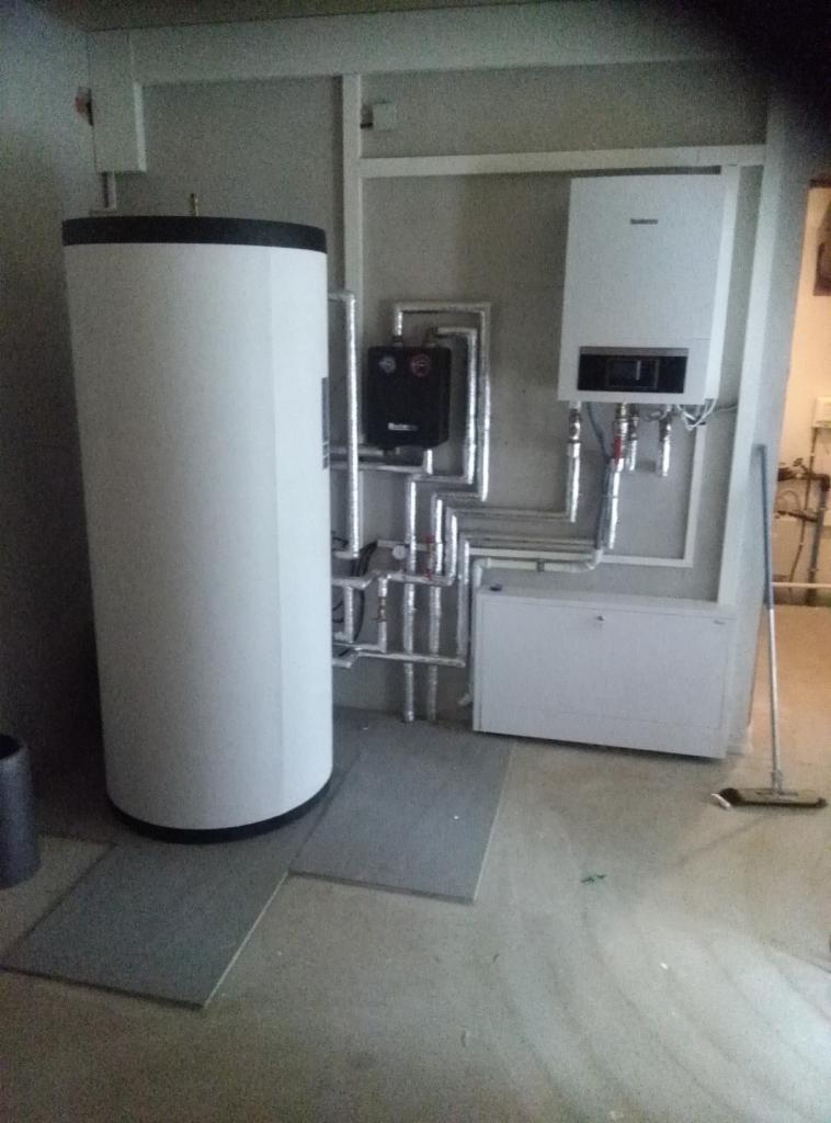 Biały, pionowy zbiornik na wodę, wiszący na ścianie biały piec grzewczy i rury w izolacji, zainstalowane w pomieszczeniu o szarych ścianach i podłodze, z mopem opartym o ścianę po prawej stronie.