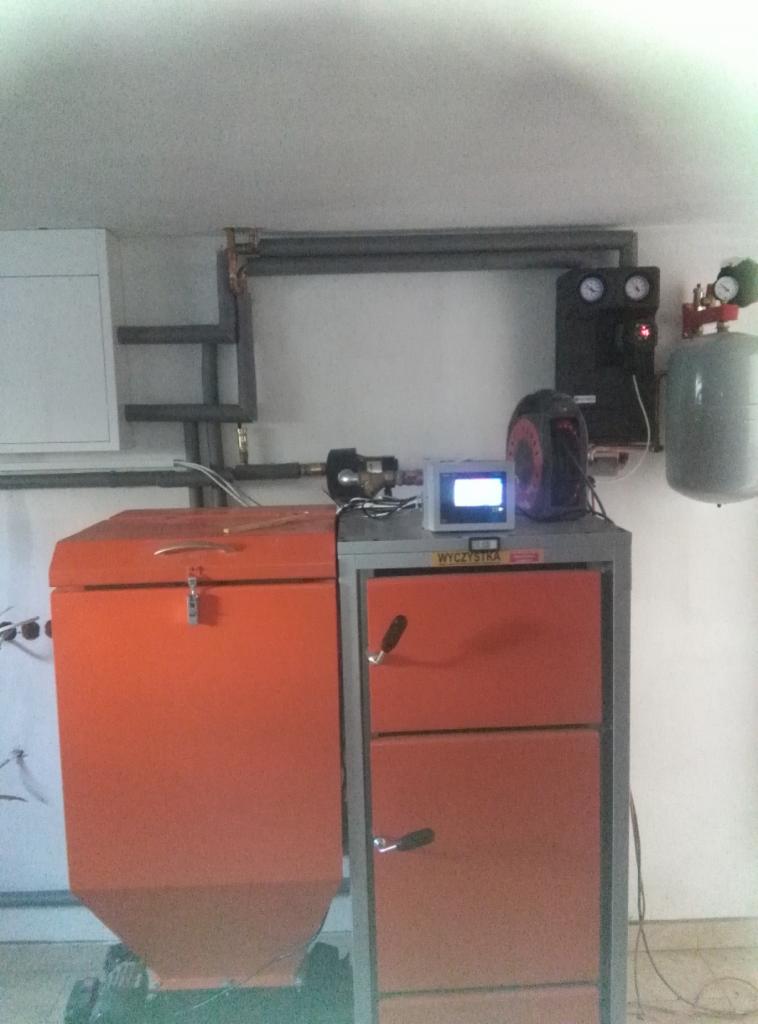 Pomarańczowy piec na paliwo stałe z podajnikiem i elektronicznym sterownikiem w kotłowni w miejscowości Biała druga, z widocznymi elementami instalacji grzewczej.