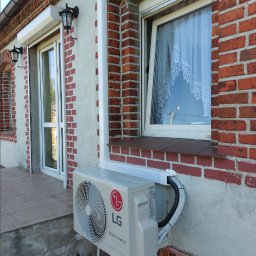 BIO - KLIM - Zewnętrzna jednostka klimatyzacji LG Dual Inverter zamontowana na ceglanej ścianie domu z białymi ramami okiennymi i ozdobną firanką w oknie.