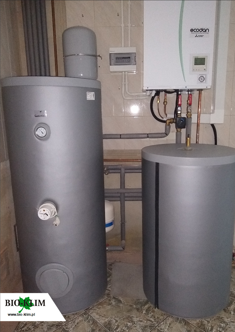 Szare zbiorniki akumulacyjne ciepłej wody użytkowej marki Galmet i Ecodan, z widocznymi rurami instalacyjnymi i sterownikiem elektrycznym na ścianie z beżowych płytek.
