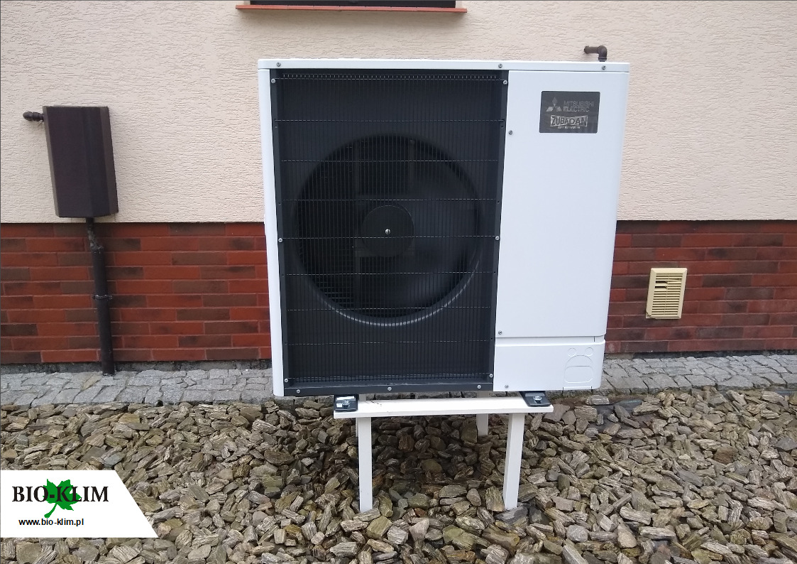 Biała pompa ciepła Mitsubishi Zubadan zamontowana na podwyższeniu na tle beżowej ściany z ceglaną podmurówką i brązowym odpływem.