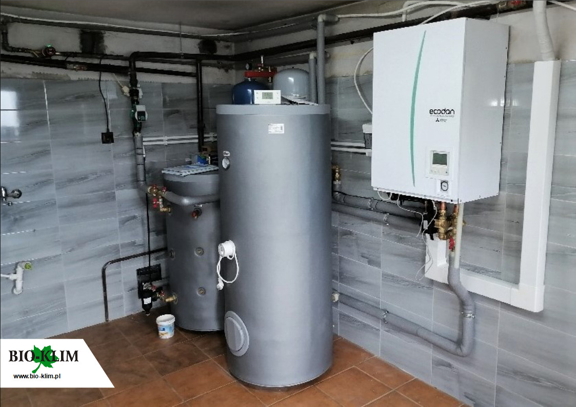 Zestaw dwóch szarych, pionowych zbiorników akumulacyjnych ciepła oraz biała jednostka zewnętrzna z zielonym akcentem, marki Ecodan Mitsubishi Electric, zamontowane w kotłowni z szaro-białymi...