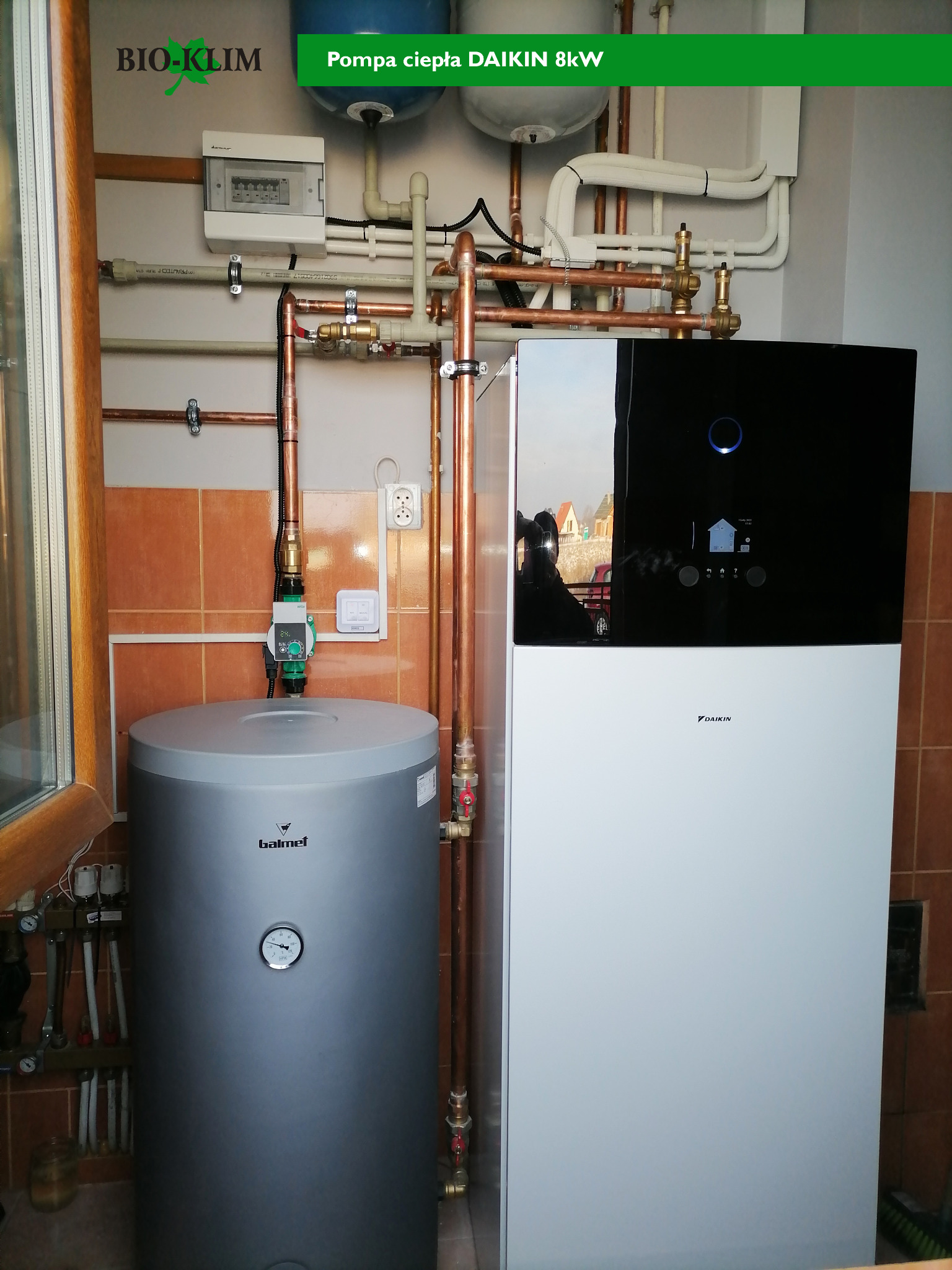 Szara pionowa instalacja zbiornika Galmet i biało-czarna Daikin 8kW, z miedzianymi rurami, zamontowane na tle brązowych płytek ceramicznych. Widoczna skrzynka elektryczna i okno.
