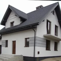 Biały dom z ciemnoszarą dachówką, elewacją z szarymi akcentami i brązowymi oknami, widok z zewnątrz z betonowymi schodami wejściowymi.