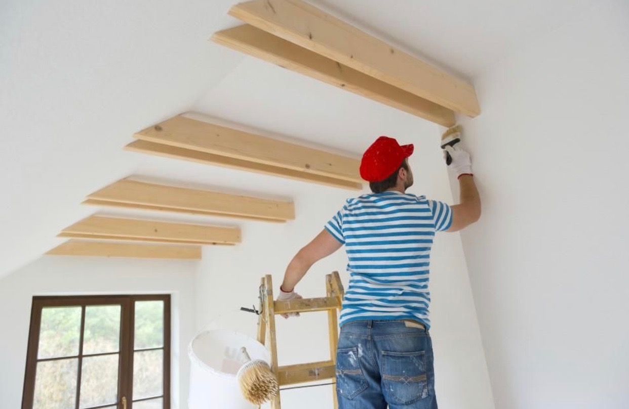 Malarz w czerwonej czapce na drabinie maluje białą ścianę z drewnianymi belkami pod sufitem, widoczny kubeł z farbą i pędzel.