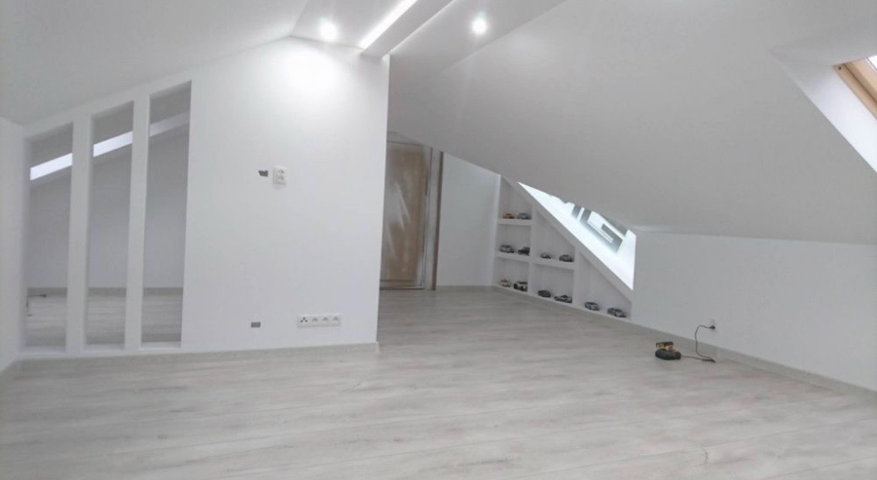 Jasne, przestronne poddasze po remoncie z białymi ścianami, drewnianą podłogą i wnękami z półkami, na których stoją małe samochody.