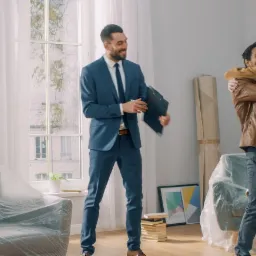 Elegancki agent nieruchomości w niebieskim garniturze wręcza klucze szczęśliwej parze w nowym, jasnym mieszkaniu z dużym oknem, gdzie meble są jeszcze zabezpieczone folią.