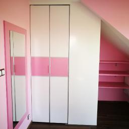 AB Transport - Biała szafa przesuwna z różowym pasem na środku, dopasowana do różowych ścian pokoju na poddaszu, obok lustro w różowej ramie i widoczne półki w skosie dachu.