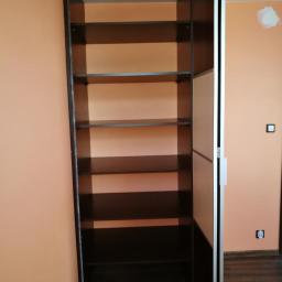 AB Transport - Wysoka, pusta szafa wnękowa z ciemnego drewna z półkami, z częściowo otwartymi drzwiami przesuwnymi, na tle pomarańczowej ściany z widocznym gniazdkiem.