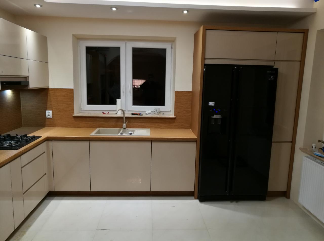 Jasna kuchnia z beżowymi frontami szafek, drewnianym blatem i czarną lodówką side-by-side, widoczne narzędzia na parapecie sugerują montaż.