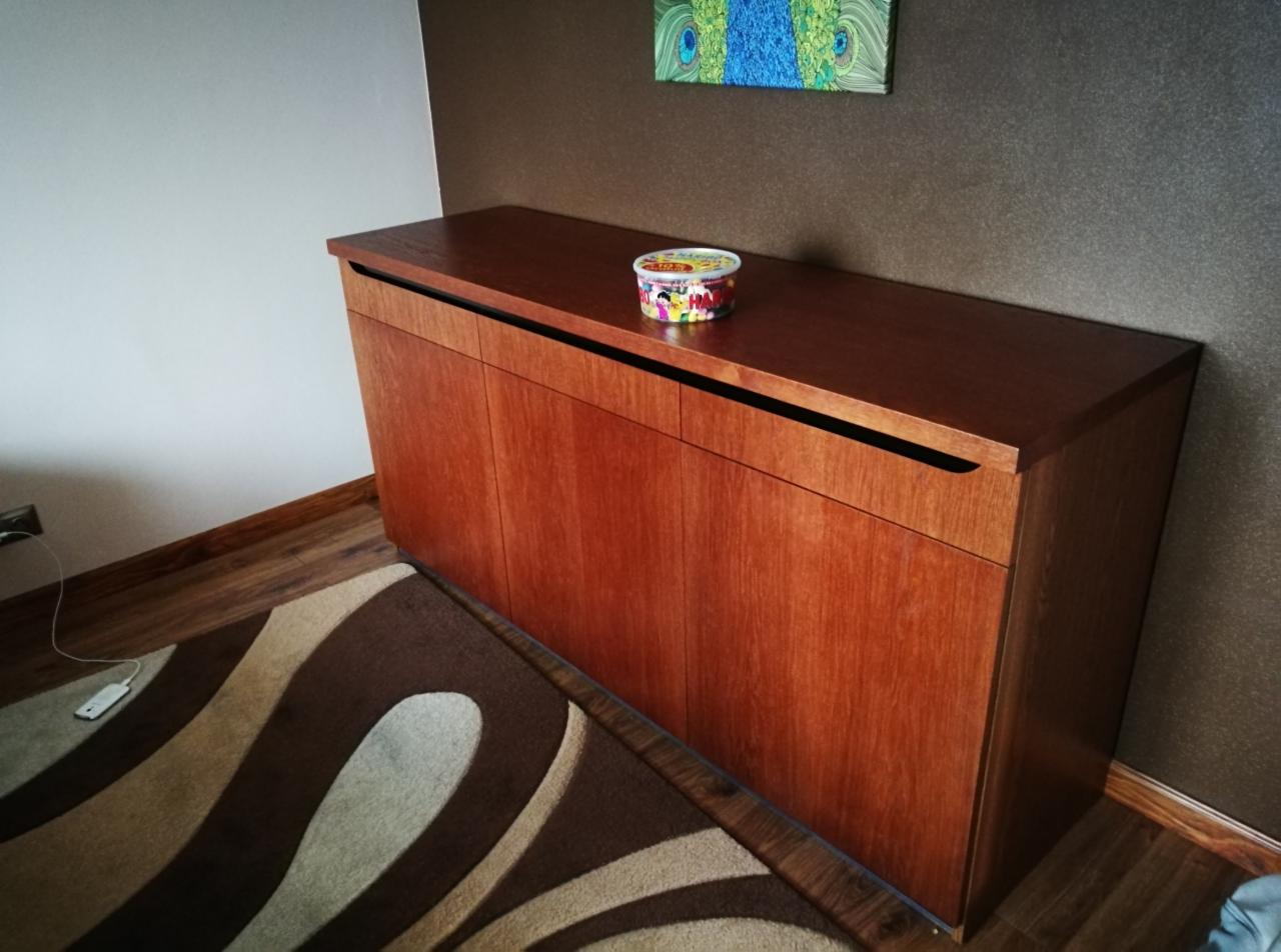 Drewniana komoda z czterema drzwiczkami z ukrytymi uchwytami, na blacie kolorowa puszka, w tle brązowa ściana z obrazem pawiego oka, na podłodze wzorzysty dywan.