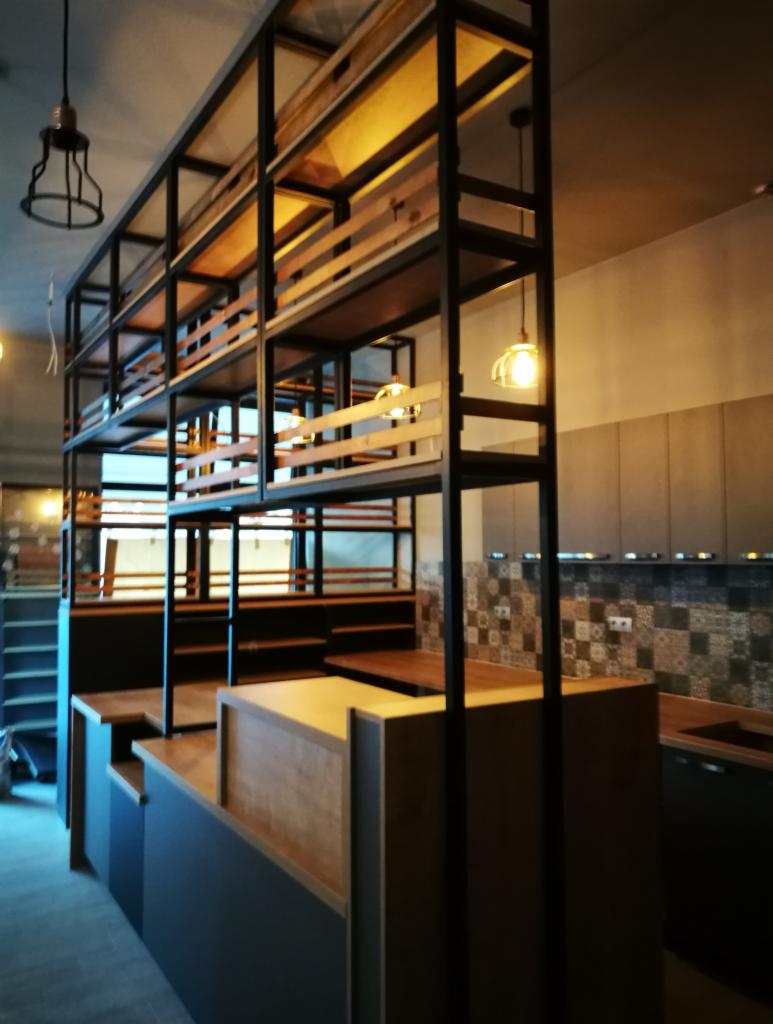 Wysoka, otwarta konstrukcja regałowa z metalu i drewna, oddzielająca przestrzeń kuchenną z mozaikową ścianą, widoczne oświetlenie wiszące.