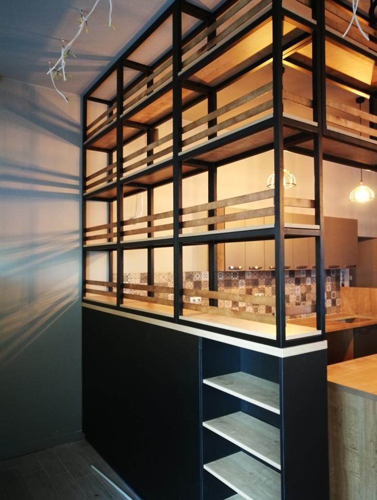 Czarno-drewniana ażurowa ścianka działowa z półkami, oddzielająca kuchnię, z widocznymi elementami zabudowy kuchennej w tle i wiszącymi lampami.