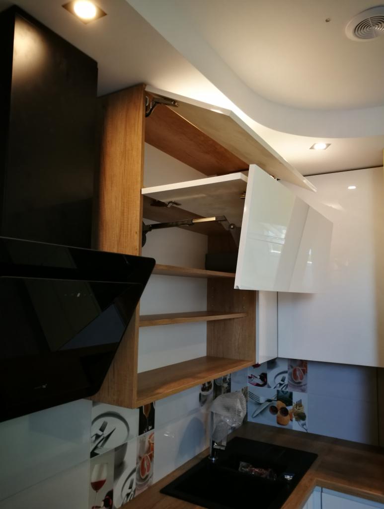 Otwarta szafka kuchenna z drewnianymi półkami i białymi frontami, widoczny mechanizm podnoszenia frontu, czarny okap i płytki z motywami kulinarnymi.