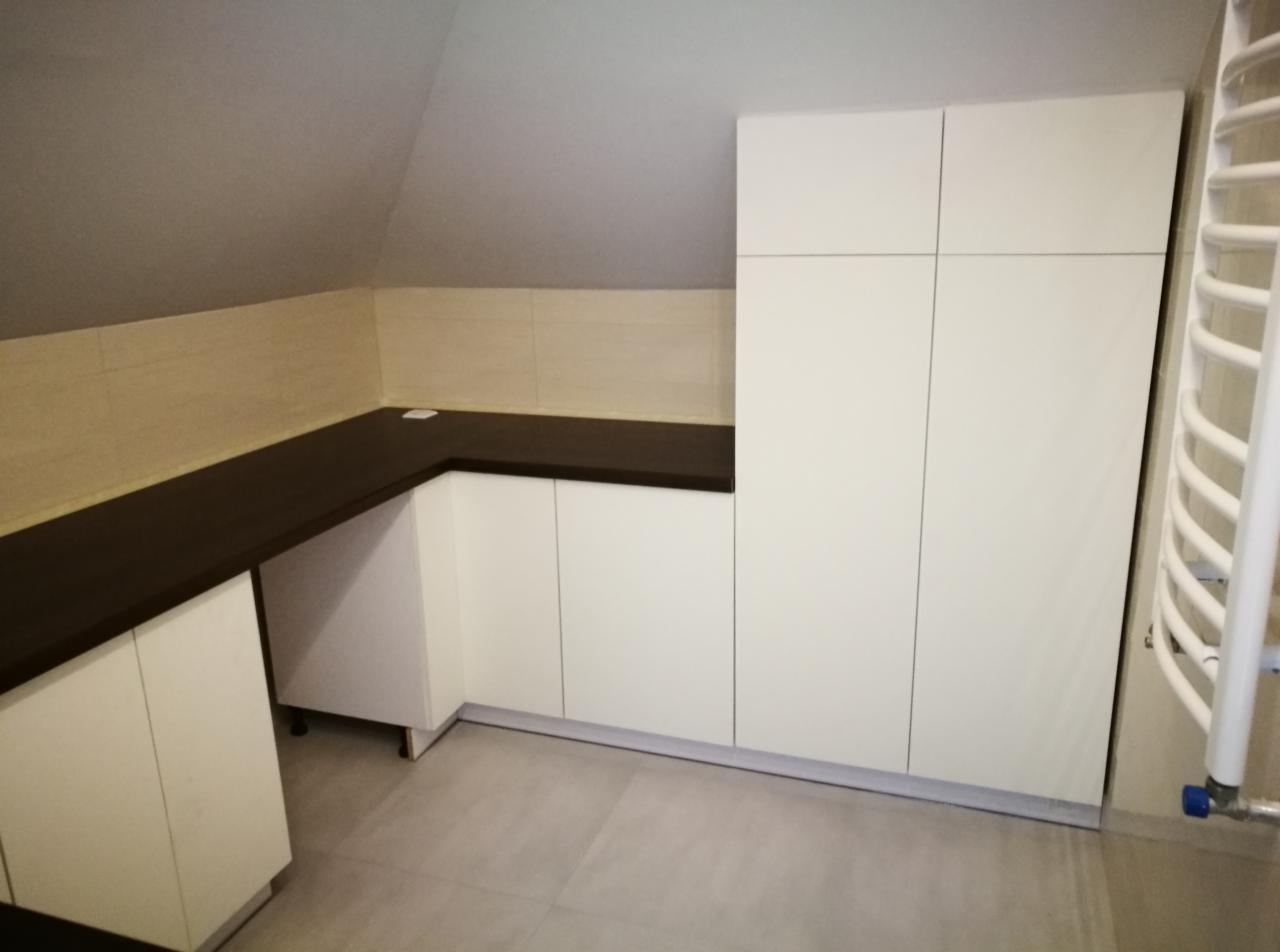 Białe szafki kuchenne pod skosem dachu z ciemnobrązowym blatem, widoczny grzejnik drabinkowy.