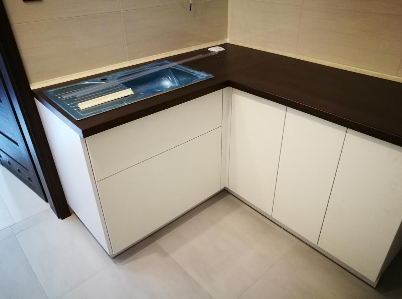 Nowoczesna kuchnia narożna z białymi frontami i ciemnobrązowym blatem, zlew zabezpieczony folią ochronną, widoczne płytki podłogowe w odcieniach szarości.