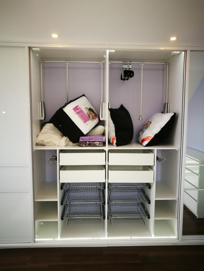 Wnętrze białej szafy przesuwnej z półkami, szufladami i koszami drucianymi, wypełnionej pościelą i poduszkami, oświetlone od dołu listwą LED.
