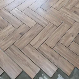 Ułożona podłoga z drewnopodobnych płytek w jodełkę, widoczne świeże ślady zaprawy i fragment białej listwy przypodłogowej.