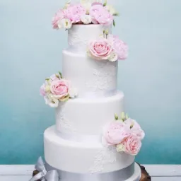 Czteropiętrowy tort weselny w stylu rustykalnym, dekorowany białym lukrem, koronkowymi zdobieniami, różowymi kwiatami i srebrną wstążką, umieszczony na drewnianym pniu z imionami Kinga i Daniel...