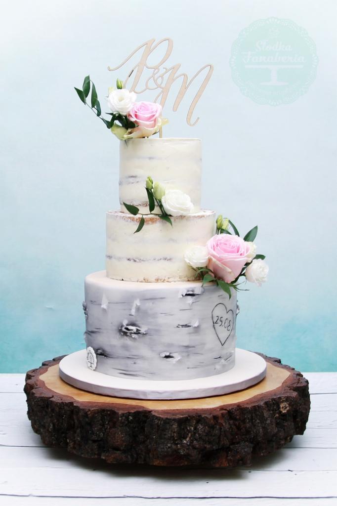 Trzypiętrowy tort weselny w stylu rustykalnym, zdobiony kremem, świeżymi kwiatami i topperem z inicjałami na drewnianym postumencie imitującym pień drzewa.