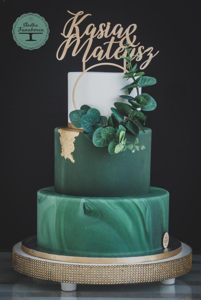 Trzypiętrowy tort weselny w odcieniach zieleni z marmurkowym wzorem na dolnej warstwie, złotymi akcentami i drewnianym topperem z imionami Kasia i Mateusz, udekorowany zielonymi listkami.