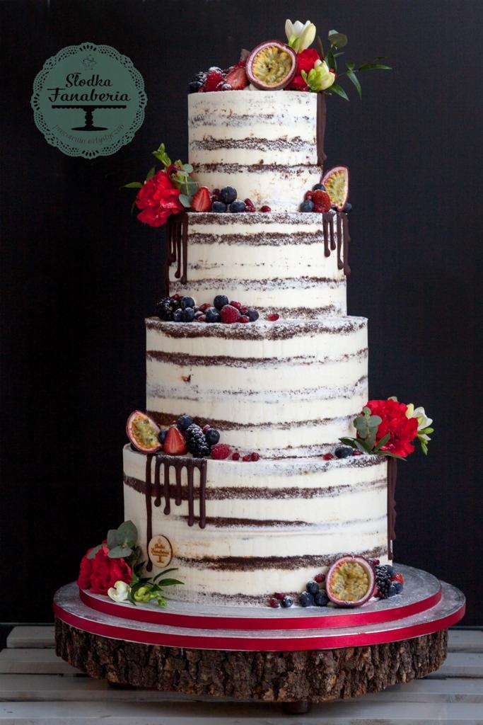 Czteropiętrowy tort 'naked cake' z kremem, owocami leśnymi, marakują, czerwonymi kwiatami i polewą czekoladową na drewnianej podstawie z czerwoną wstążką, na ciemnym tle.