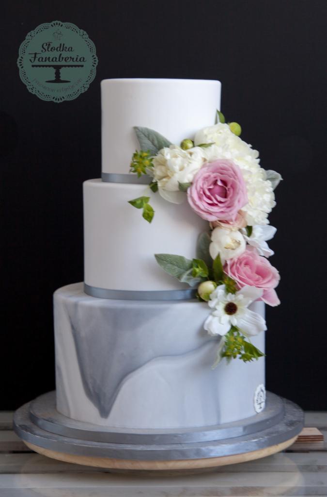 Trzypiętrowy tort weselny z marmurkowym wykończeniem dolnej warstwy i dekoracją z różowych i białych kwiatów, ozdobiony srebrną wstążką.