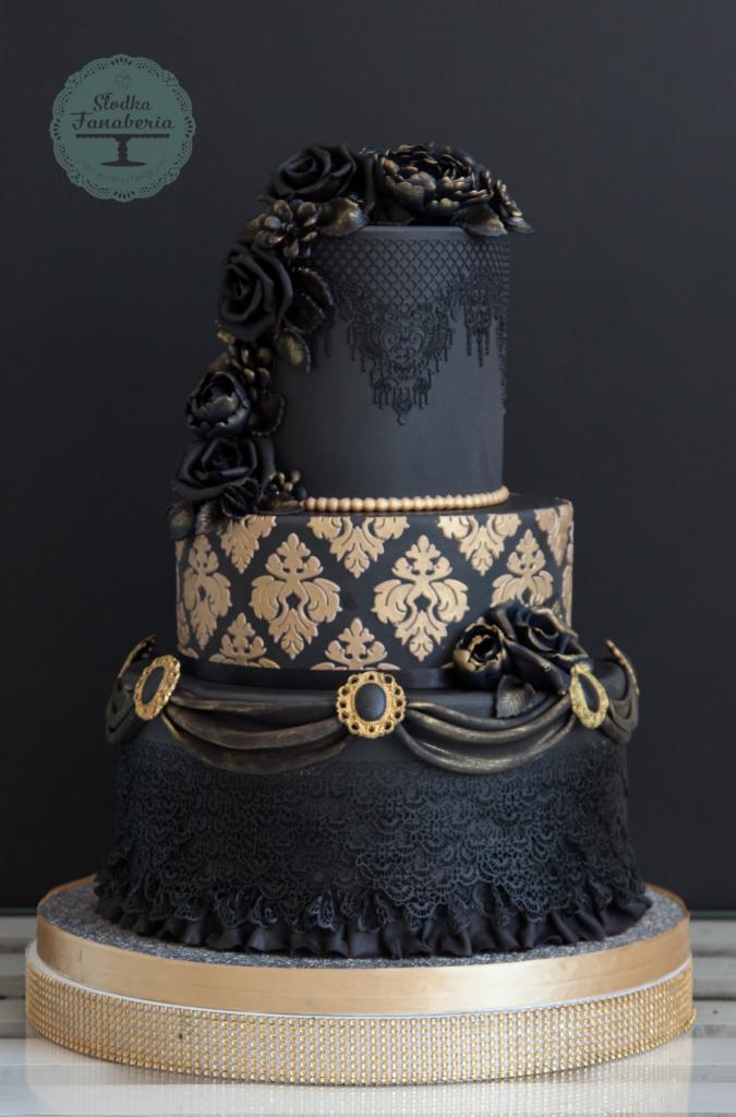 Elegancki, trzypiętrowy tort w czarno-złotej kolorystyce, zdobiony koronką, wzorami damaskowymi, perłami i kwiatami z lukru, umieszczony na złotej podstawie.