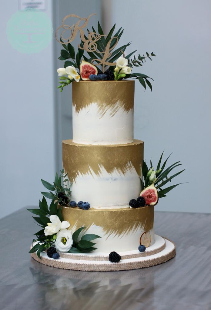 Trzypiętrowy tort weselny z efektem pociągnięć pędzlem w kolorze złota, udekorowany figami, jeżynami, borówkami i białymi kwiatami, zwieńczony drewnianym topperem z inicjałami.