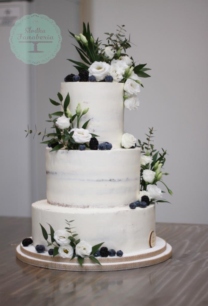 Trzypiętrowy tort weselny w stylu rustykalnym, udekorowany białymi kwiatami, jagodami i zielonymi liśćmi, umieszczony na podstawie z jutowym obramowaniem.