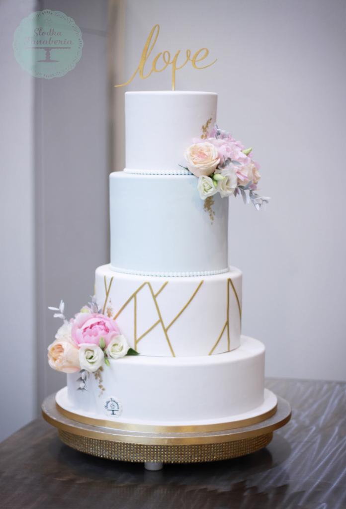 Czteropiętrowy tort weselny w białej polewie, zdobiony złotymi geometrycznymi wzorami, świeżymi kwiatami w odcieniach różu i bieli oraz złotym napisem 'love' na szczycie, umieszczony na złotej...