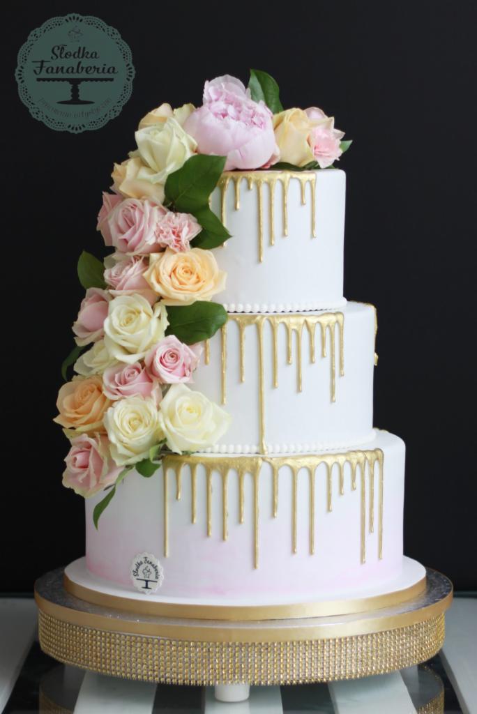 Trzypiętrowy tort weselny z białym lukrem, złotymi polewami i dekoracją z różowych i białych róż, umieszczony na złotej paterze.
