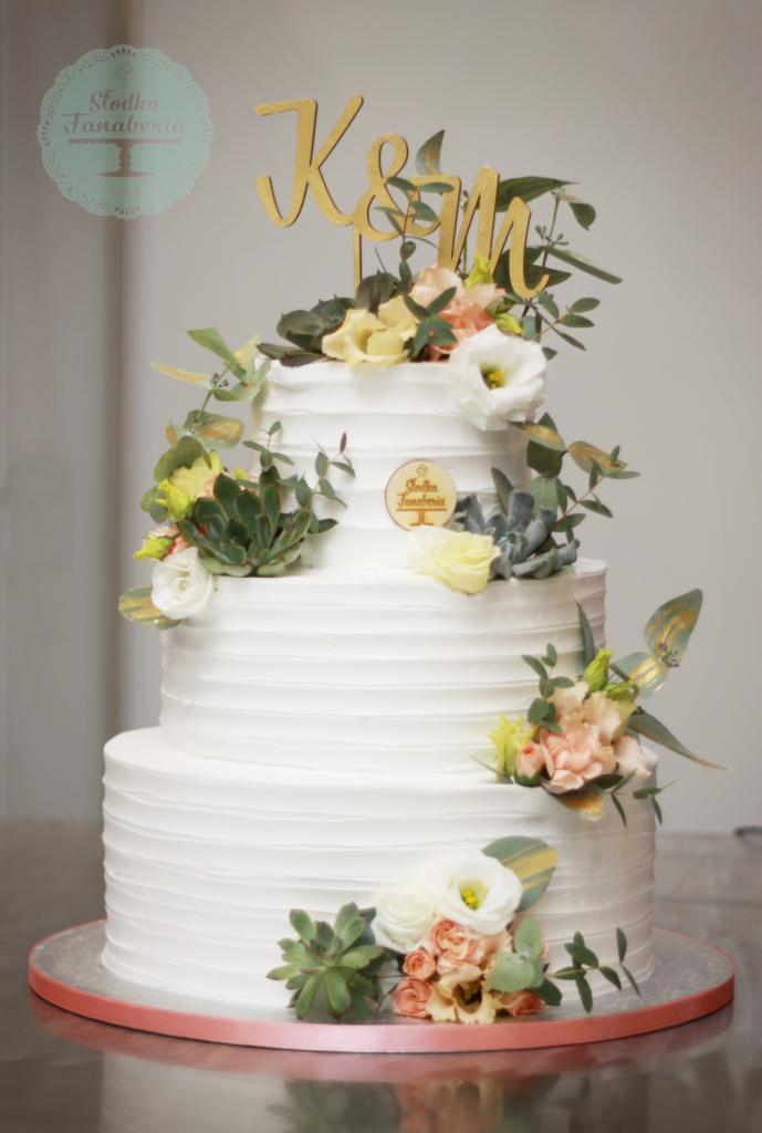 Trzypiętrowy tort weselny z białym kremem, dekorowany sukulentami, białymi i pastelowymi kwiatami oraz złotym topperem z inicjałami K&M, umieszczony na srebrnej paterze.