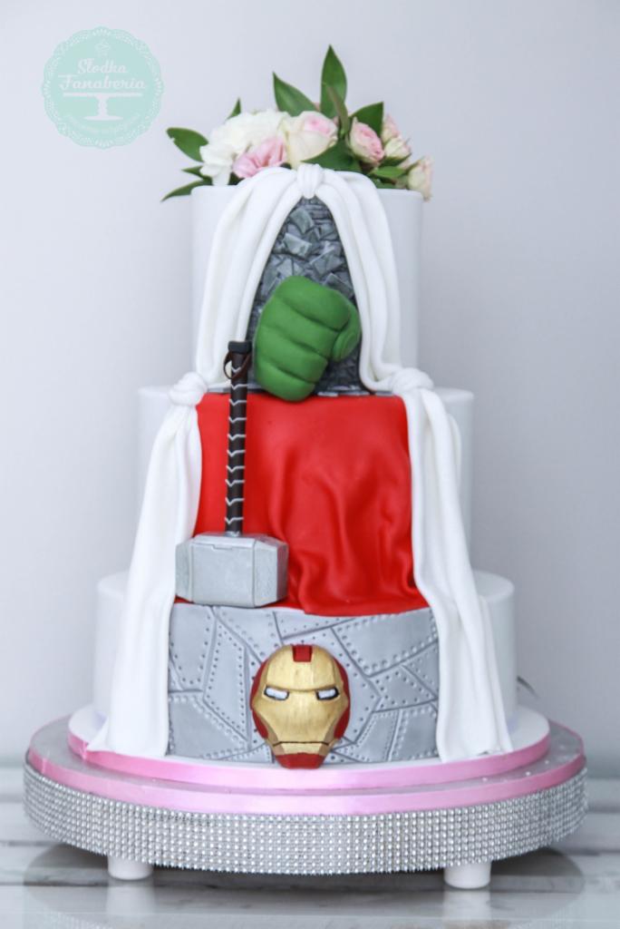 Trzypiętrowy tort z dekoracjami nawiązującymi do Avengers: rękawica Hulka, młot Thora i maska Iron Mana, całość zwieńczona bukietem kwiatów.