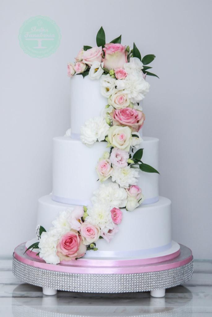 Trzypiętrowy tort weselny w minimalistycznym stylu, udekorowany kaskadą różowych i białych kwiatów oraz zielonymi liśćmi, umieszczony na srebrnej, zdobionej podstawie z różową wstążką.