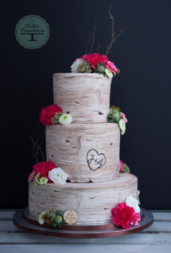 Trzypiętrowy tort weselny w stylu rustykalnym, imitujący korę brzozy, ozdobiony świeżymi, różowymi i białymi kwiatami oraz inicjałami w sercu.